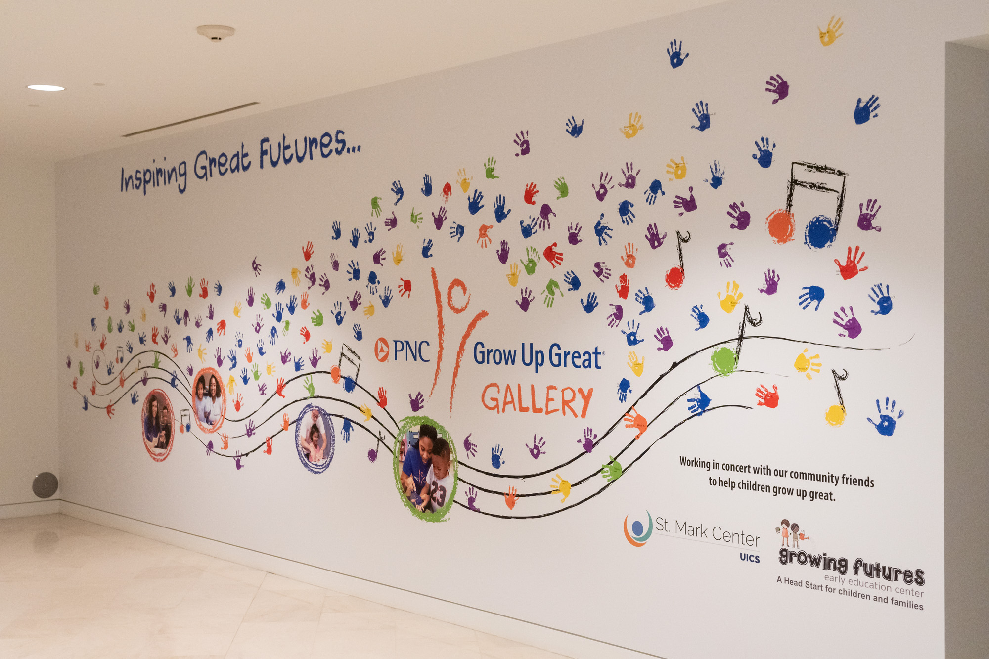 A wall mural titled "Inspiring Great Futures" features colorful handprints, musical notes, and children's photos, seamlessly integrating with the architecture and spaces of the PNC Grow Up Great Gallery in partnership with St. Mark Center.