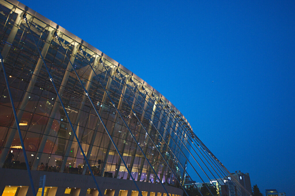 A modern glass building with curved architecture is illuminated against a deep blue evening sky. Inside lights and visible people suggest an event or gathering occurring inside.