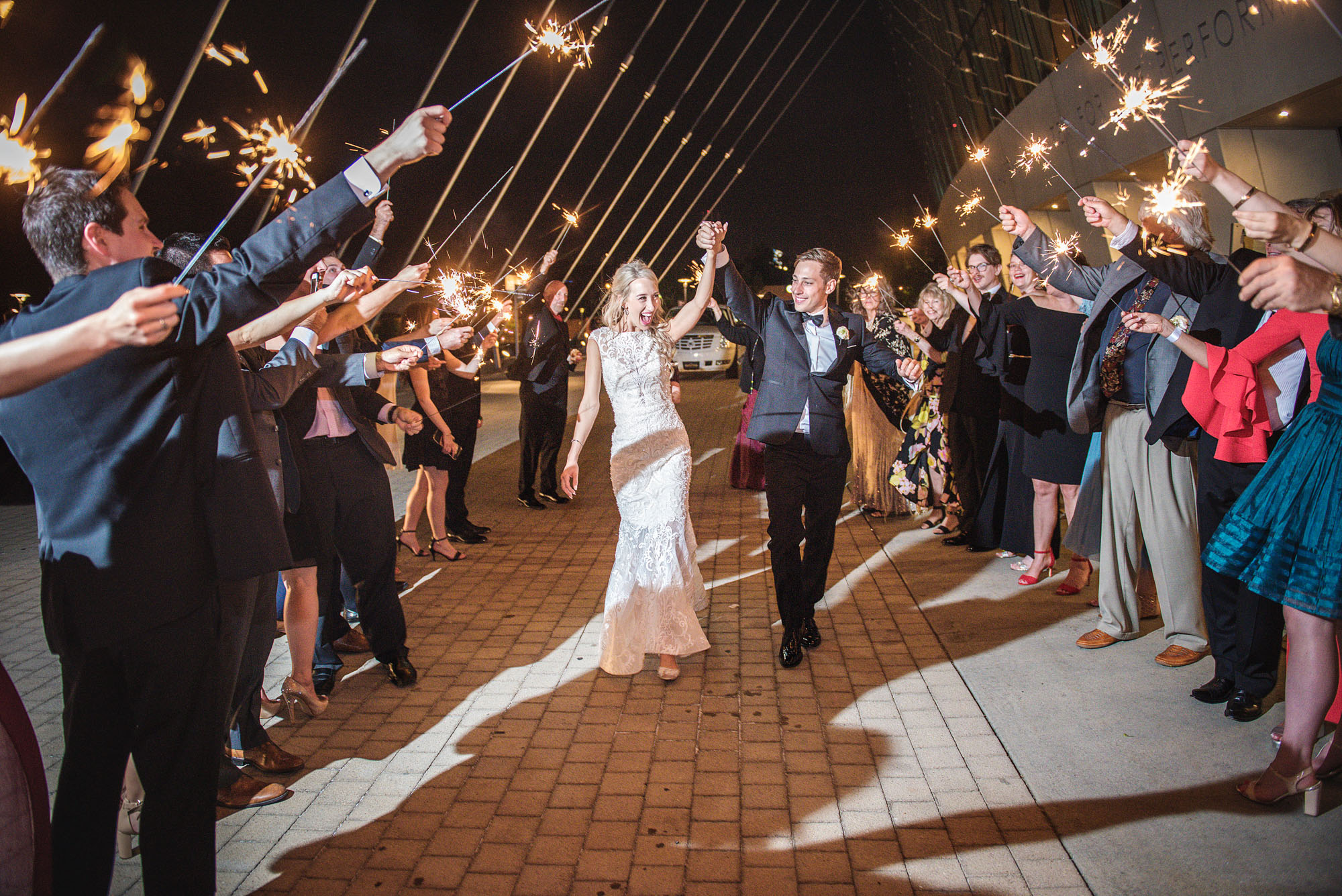 A bride and groom walk through an arch of sparklers held by a group of people at night, celebrating their wedding. The event, which could inspire those looking to rent Kauffman Center for their own special day, takes place on a brick pathway.