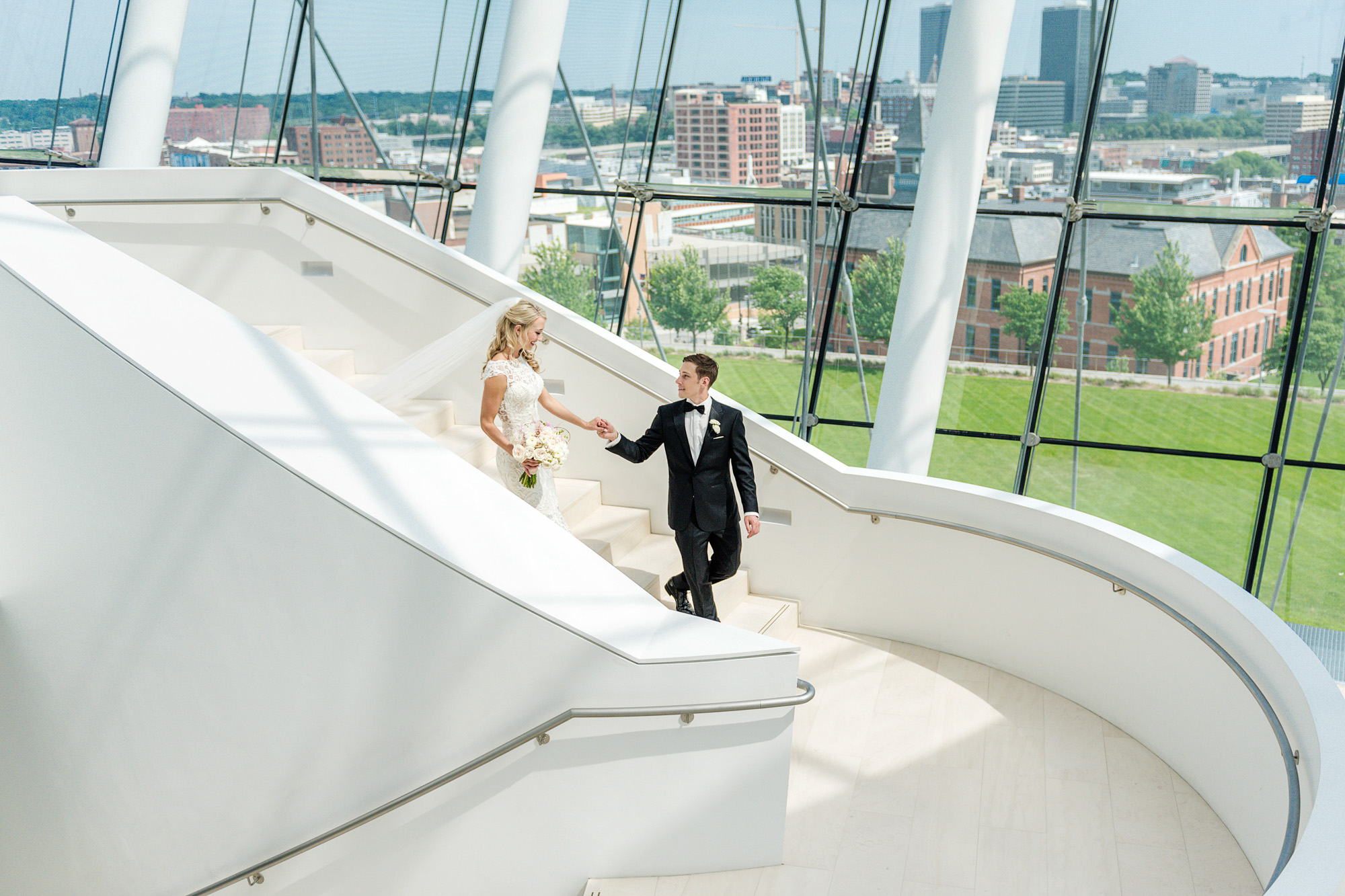 A bride and groom are walking down a curved, white staircase inside a modern building with large windows overlooking a cityscape. The groom is holding the bride's hand as she descends the stairs. For more about our venue, contact us today to make your dream wedding come true.