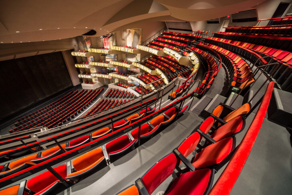 A modern theater with tiered seating, featuring red and orange seats, curved design, and intricate lighting, showcases exemplary architecture and thoughtfully designed spaces when viewed from an upper section.