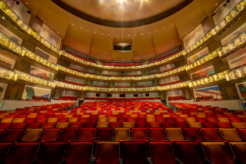 Wide-angle view of an empty, multi-tiered theater with rows of red and orange seats, ornate balconies, and a domed ceiling.