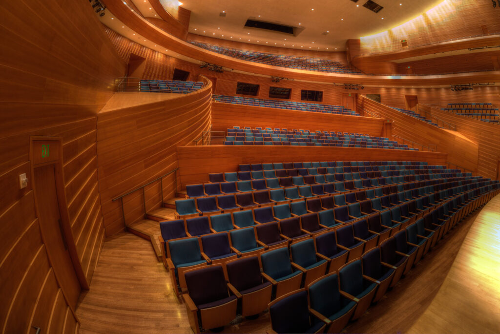 A spacious auditorium at the Kansas City Performing Arts center features wooden walls and blue cushioned seats arranged in rows, viewed from the stage. The room is empty and well-lit with tiered seating extending to the balcony.