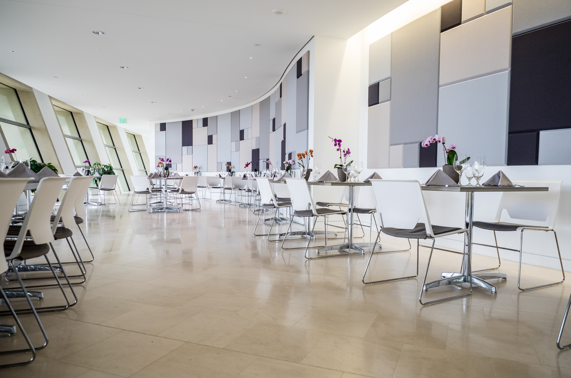 A modern dining area with white chairs and tables, set with napkins and glasses, and decorated with purple orchids, is beautifully integrated into the architecture of the space, featuring a backdrop of geometric wall panels.