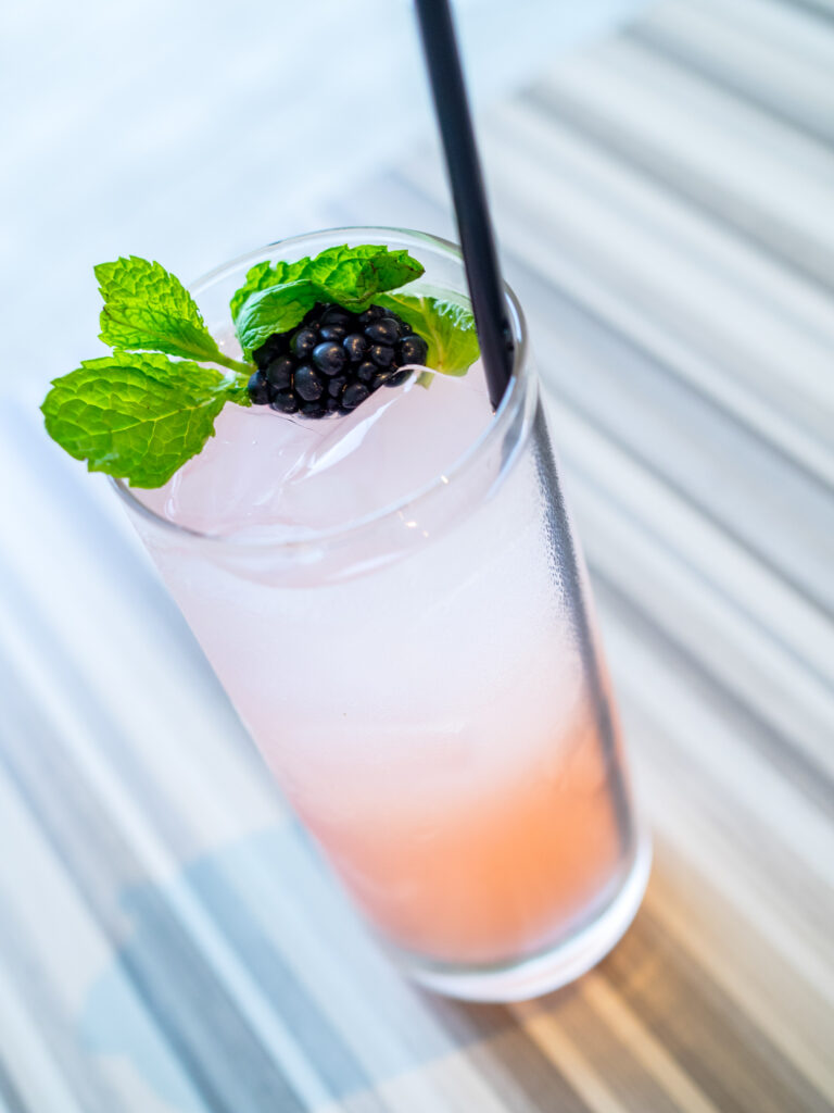 A tall glass of a pink iced beverage garnished with a blackberry, mint leaves, and a black straw on a striped surface.