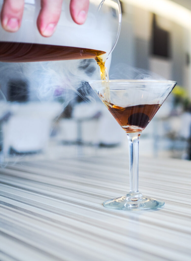 A person pours a brown liquid from a pitcher into a martini glass on a striped table. White vapor is visible around the glass.