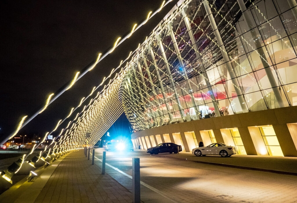 An angled exterior view of a modern glass building at night with illuminated string lights and a few parked cars.