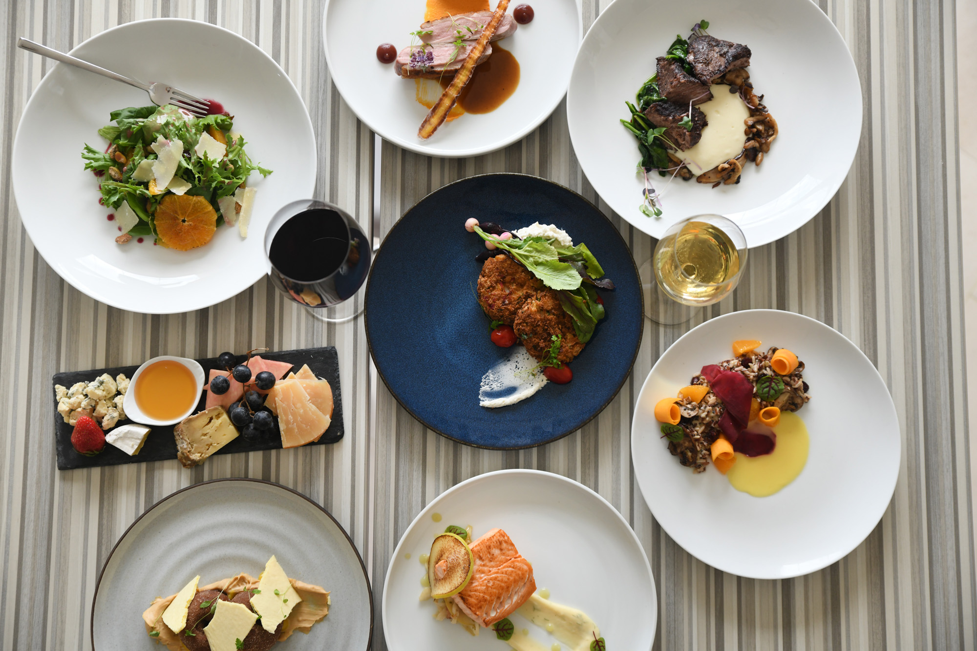 An overhead view of several plates of gourmet food, including salads, meats, cheeses, and wine, arranged on a striped table.