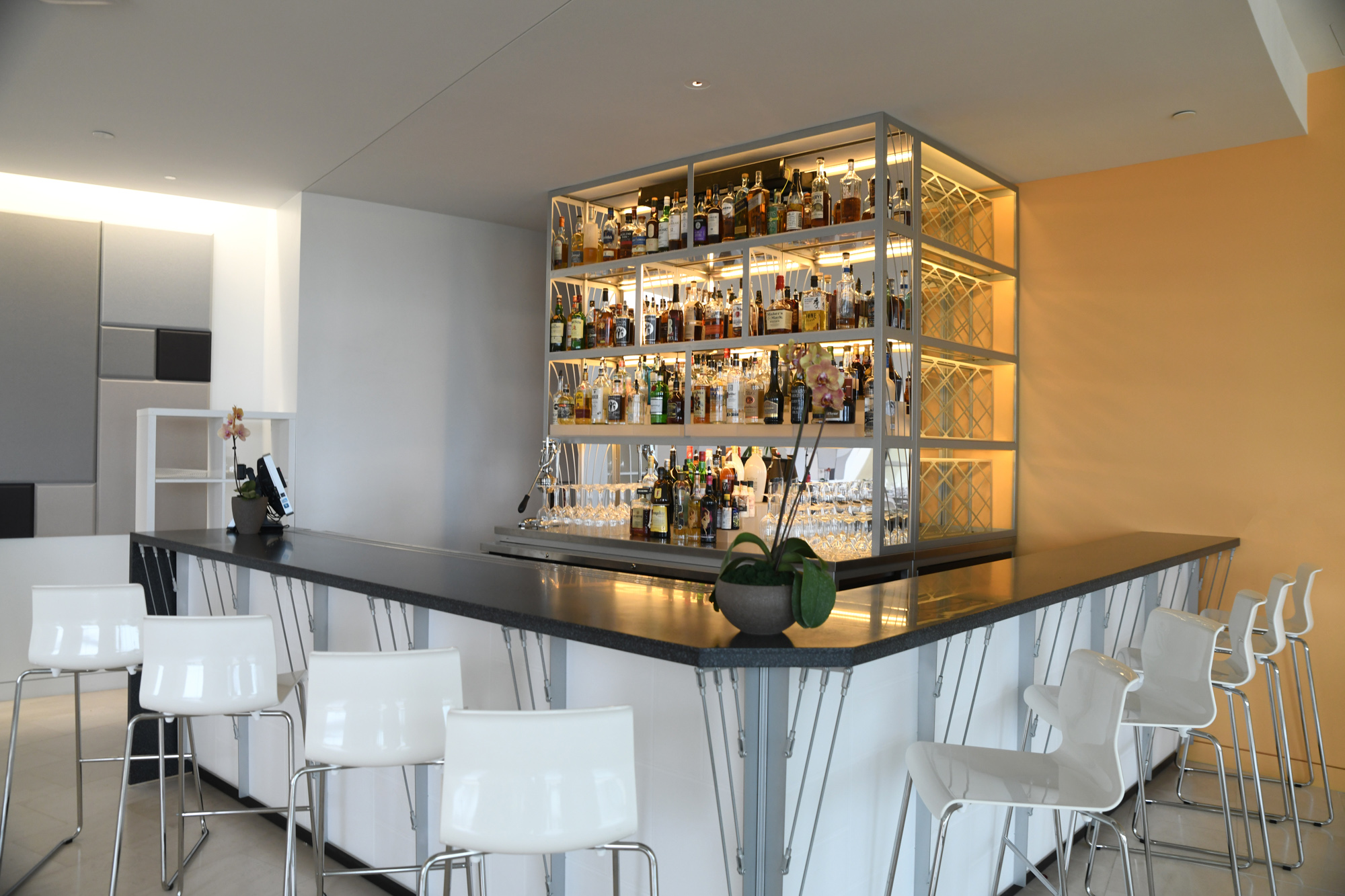 A modern bar with a sleek black countertop and white barstools arranged around it. The bar features a backlit shelf stocked with various bottles of alcohol.