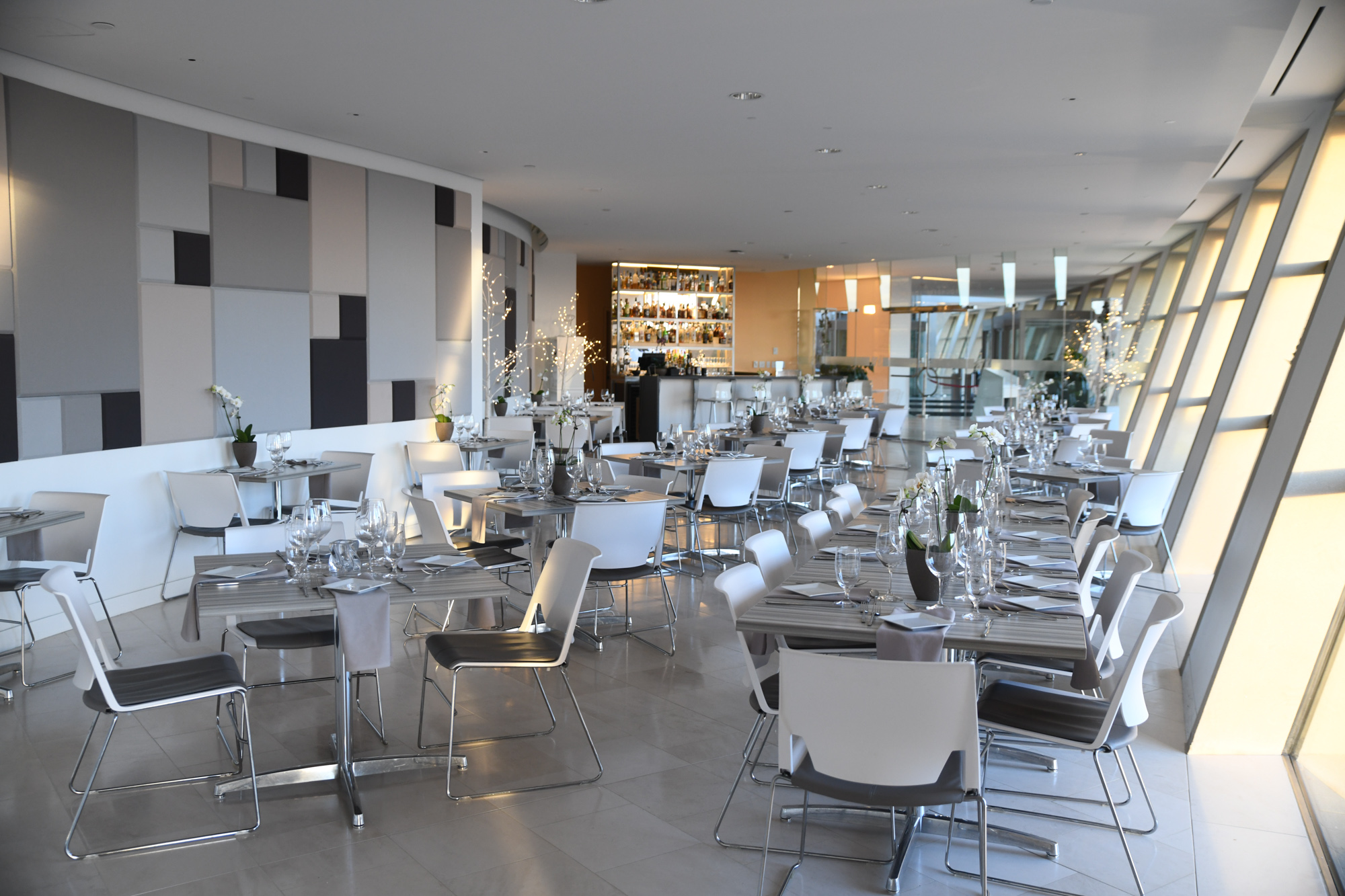 A modern restaurant interior with geometric wall design, tables set with glassware and silverware, white chairs, and a bar in the background invites diners at Kauffman Center. Large windows allow natural light to fill the space.