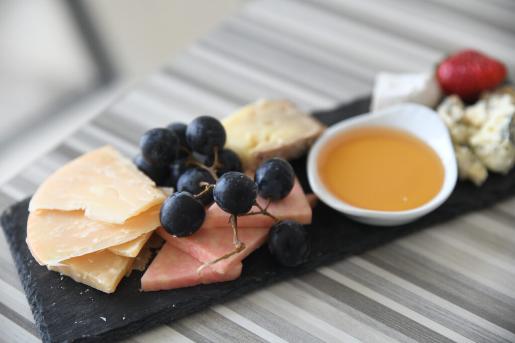 A serving of assorted cheeses, black grapes, a strawberry, and a dish of honey displayed on a black slate platter set on a striped surface.