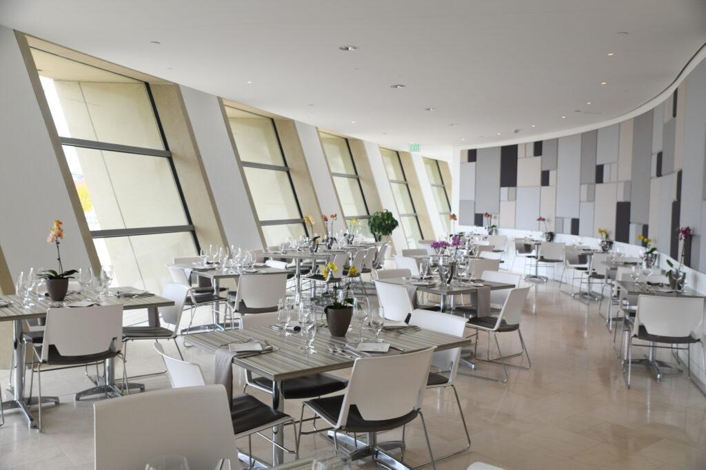 A modern restaurant interior with slanted windows, white chairs, and wooden tables set with wine glasses and cutlery. The décor includes potted orchid plants and geometric wall patterns. Perfect for dining near Kauffman Center, this space combines elegance with contemporary design.