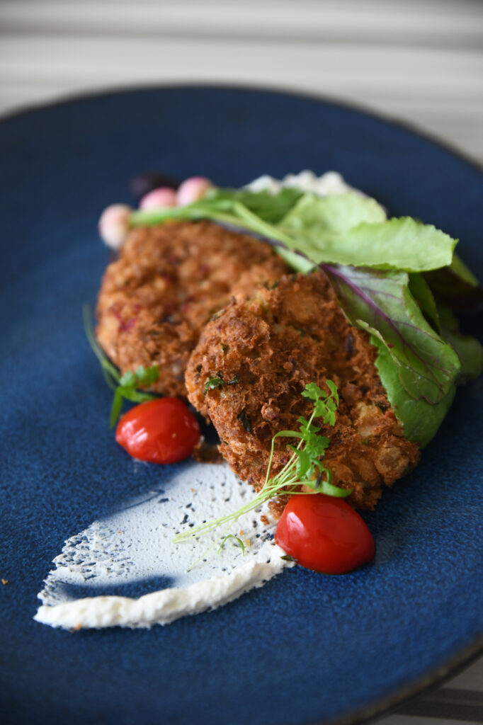 Two crispy fried pieces of food garnished with leafy greens, two cherry tomatoes, and a smear of white sauce on a blue plate.