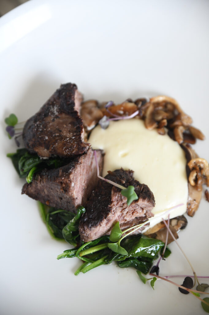 A gourmet dish featuring seared beef, sautéed spinach, mushrooms, and creamy mashed potatoes is garnished with microgreens on a white plate.