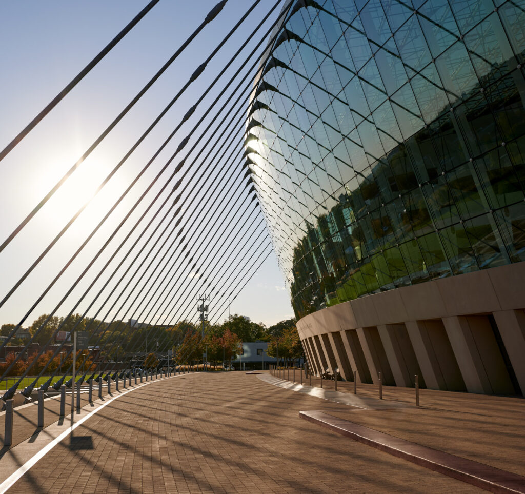 A modern building with a curved glass facade reflects sunlight. The structure is supported by numerous cables extending from the roof to the ground. A paved walkway and green area are nearby.