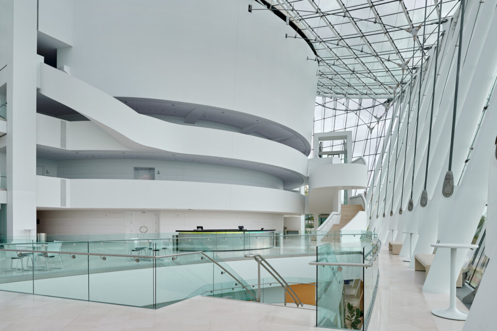 A modern, spacious indoor area with glass walls, minimalist white structures, curved balconies, and a reception desk. Stairs connect different levels and natural light floods the space.