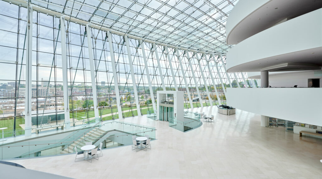 A spacious, modern atrium with large glass windows, a staircase, round tables with chairs, and a curved upper balcony.