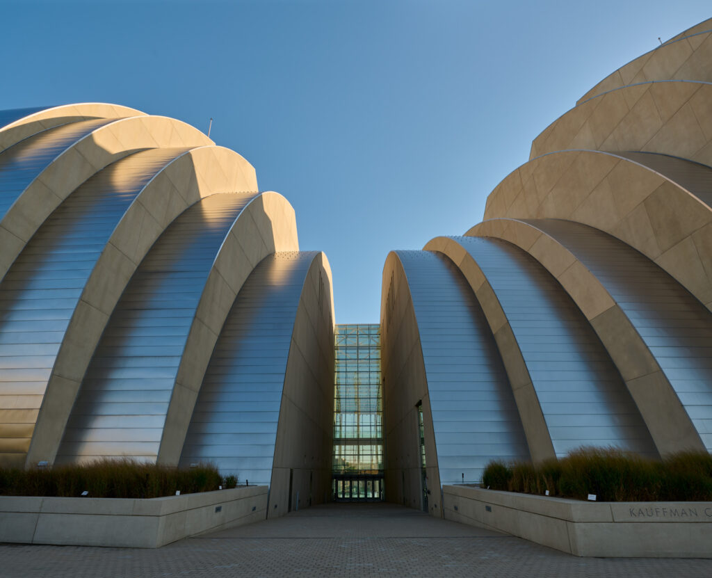 A modern building with large, curved, silver metallic panels and a glass entrance under a clear blue sky. The structure is identified as "Kauffman" based on text seen on the right.