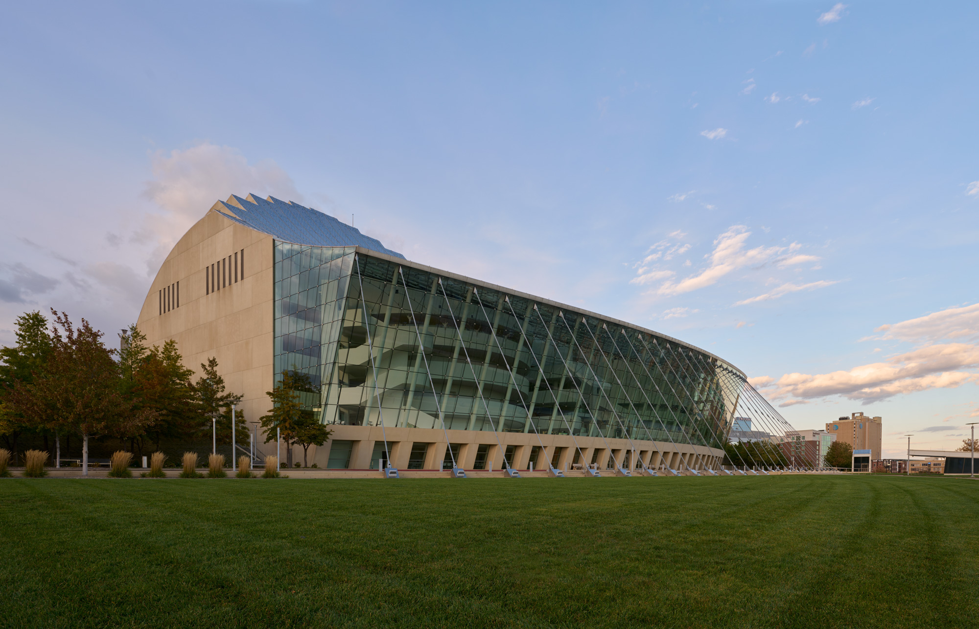 A modern building with large glass panels and angular design, reminiscent of the dynamic architecture found at Kansas City Performing Arts, sits on a green lawn under a blue sky with clouds. Trees line the left side of the structure.