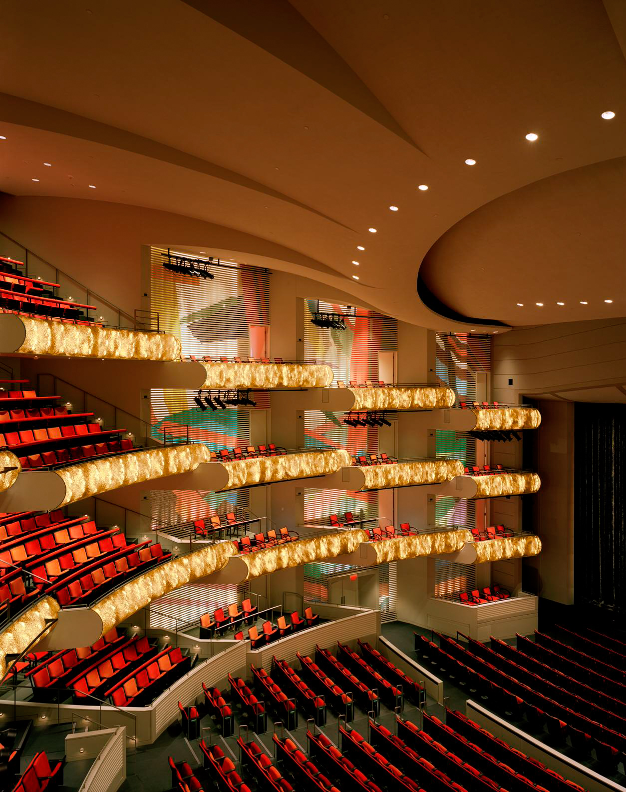 Interior view of an opera house with multiple tiers of balconies filled with empty red seats, all perfectly aligned as if placed from detailed seating maps, and illuminated by warm, ambient lighting.