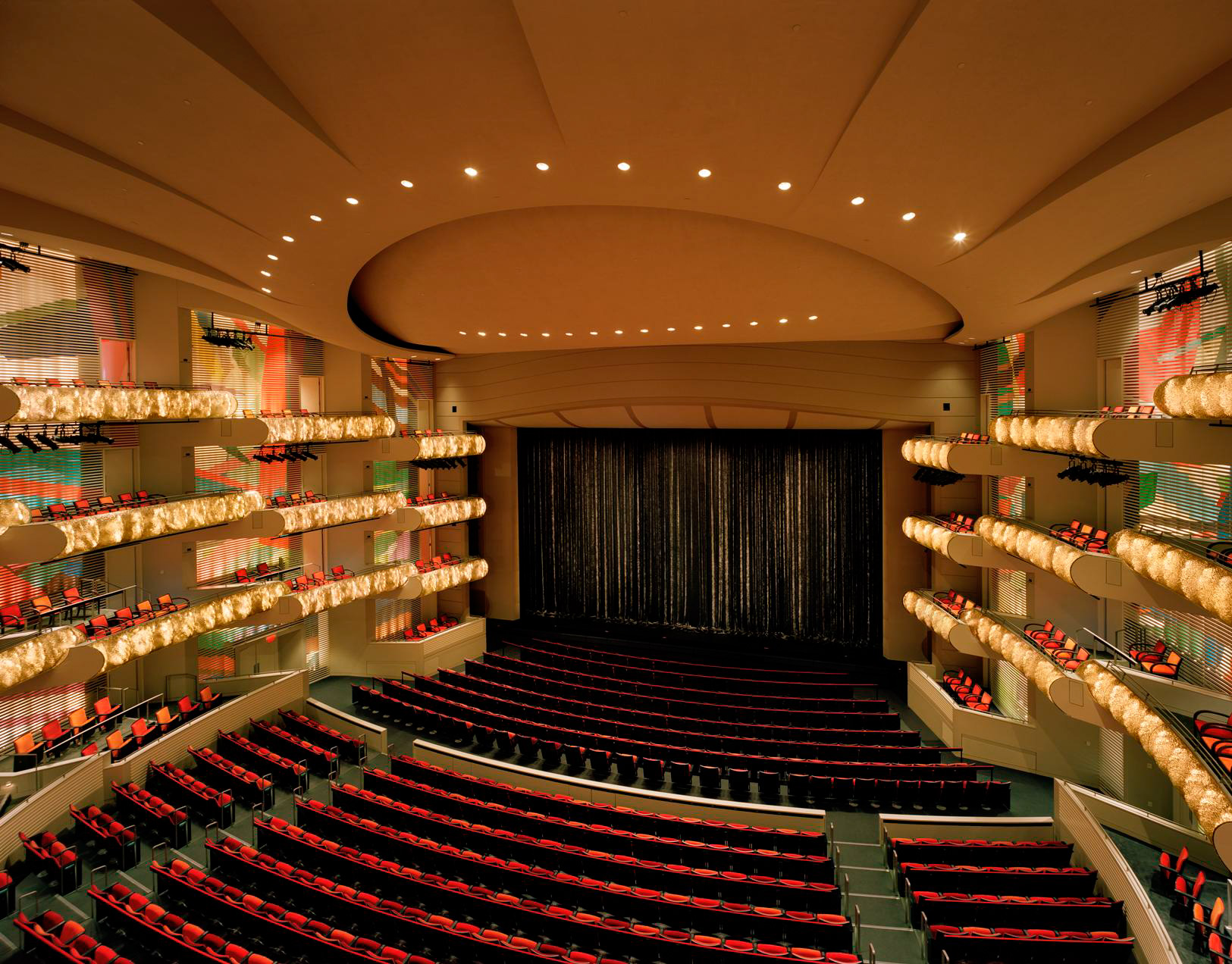 A spacious theater auditorium with red seats, multiple balconies, and a large stage with a closed curtain, the Kauffman Center Presents venue epitomizes modern elegance. The ceiling features an exquisite lighting design that enhances the ambiance for every performance.