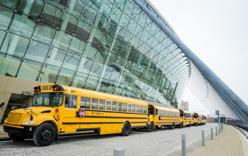 Yellow school buses are parked in a row outside the modern glass Performing Arts Center in Kansas City, which features a curved architecture. The buses are marked with school and route information. The area is paved, and the sky appears overcast.