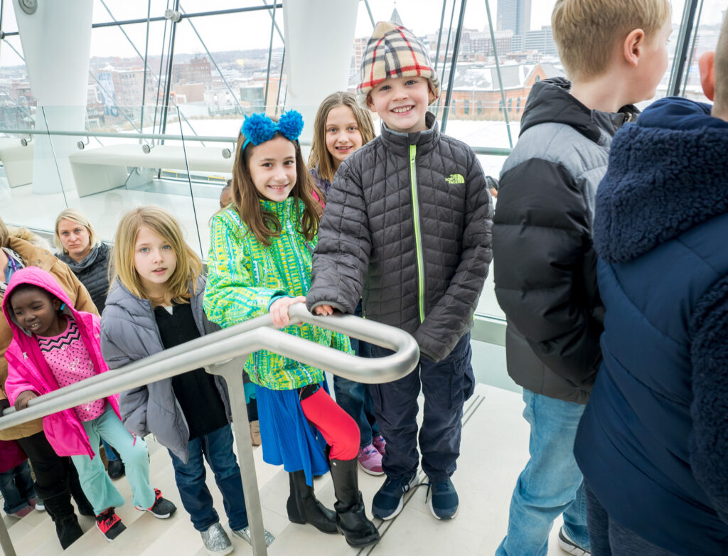 Children in winter clothing, including jackets and hats, stand in line and smile on a staircase inside a building with large windows, showcasing the joy inspired by Our Impact for the Performing Arts.