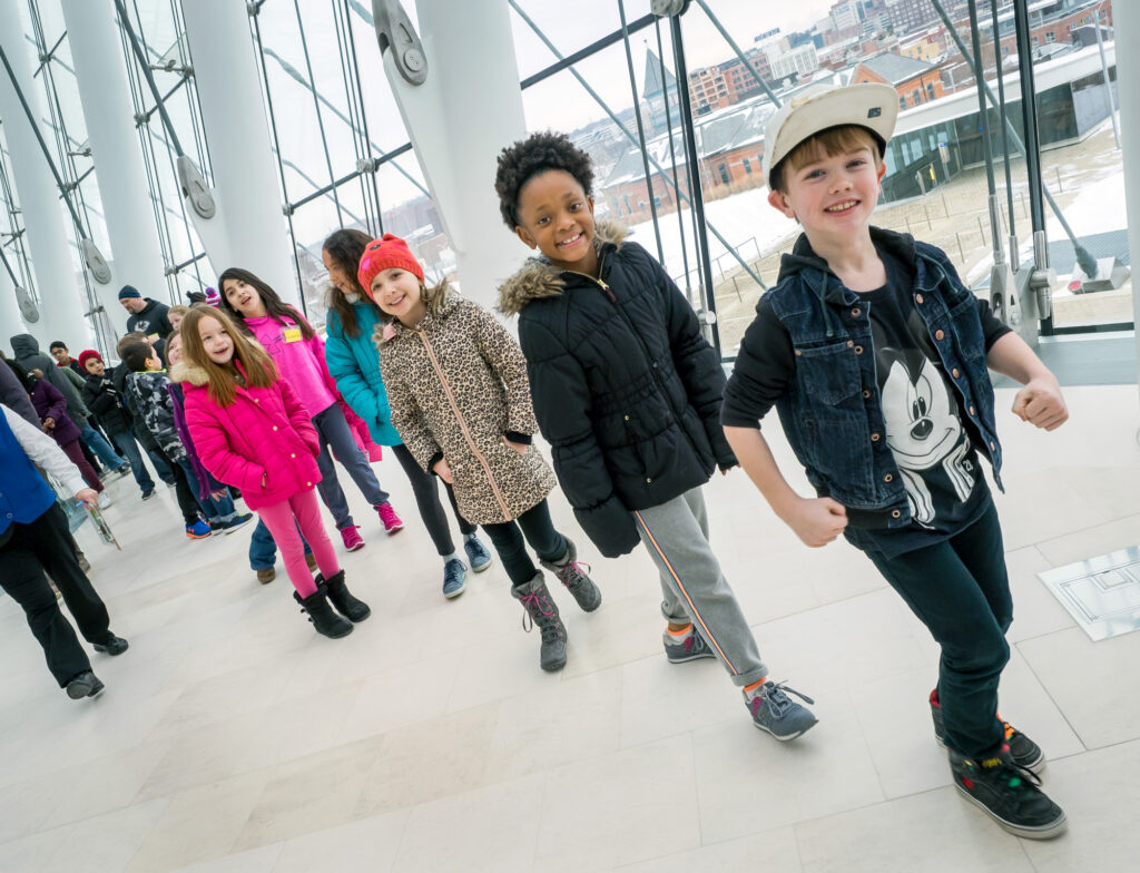 A group of children, dressed in winter clothes, walk indoors in a line with large windows showcasing a snowy cityscape. They pass by a vibrant Children’s Handprint Wall, their colorful imprints standing out against the wintry backdrop.