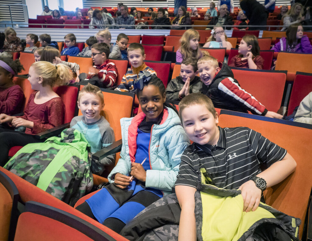 Children with student tickets sit in an auditorium, smiling and interacting with each other while waiting for the event to start.