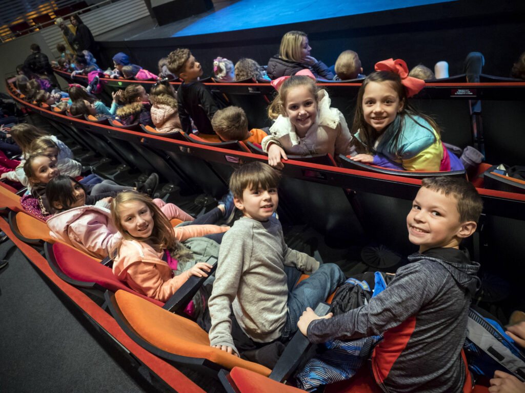 A group of children on a field trip in Kansas City sit in a theater, facing forward, while children in the background do the same. Some children are smiling at the camera.