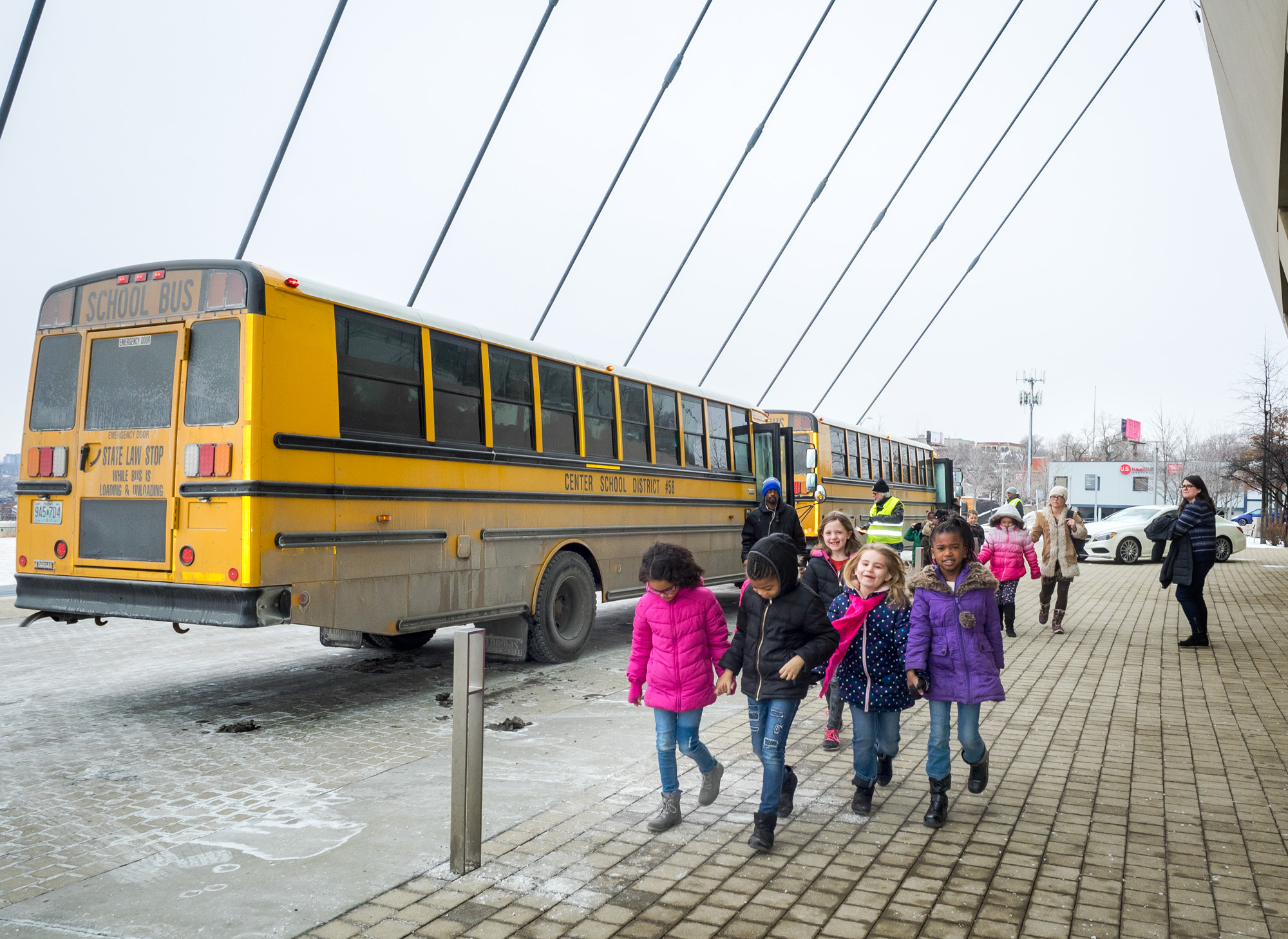 Children in winter clothing walk on a paved area near a yellow school bus, clutching their student tickets. Other school buses are parked in the background.