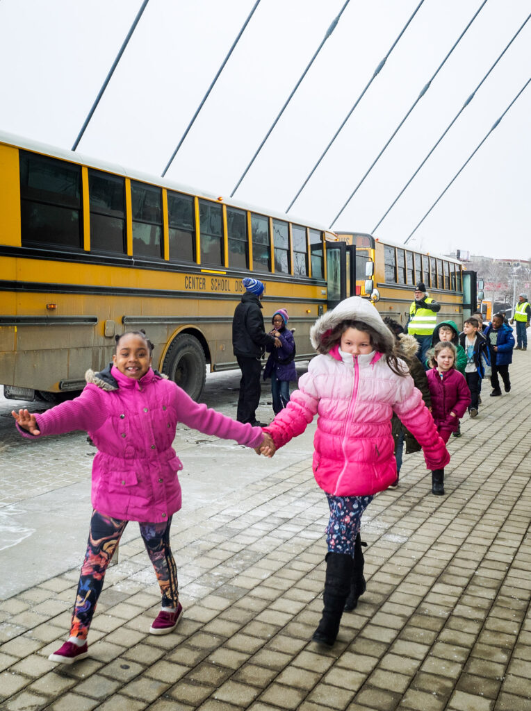 A group of children, dressed in winter clothes, hold hands and walk on a paved path beside school buses. Adults and safety officers, supported by the Kauffman Center Bus Transportation Fund, oversee their movement.