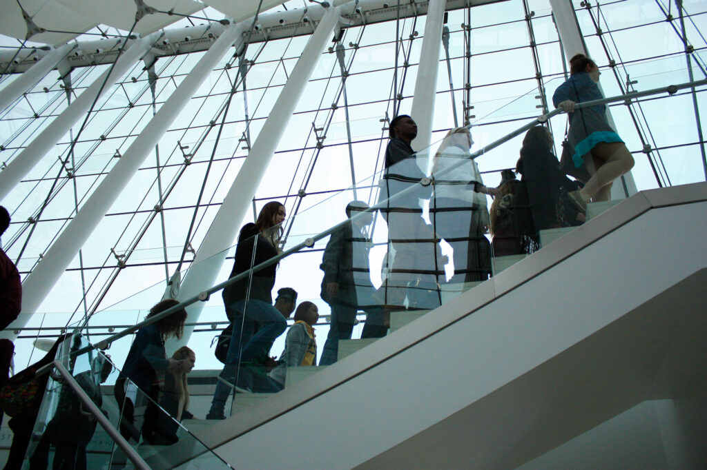 People walking up a glass-enclosed staircase, with large windows in the background letting in natural light. Request a tour to experience this architectural marvel firsthand.