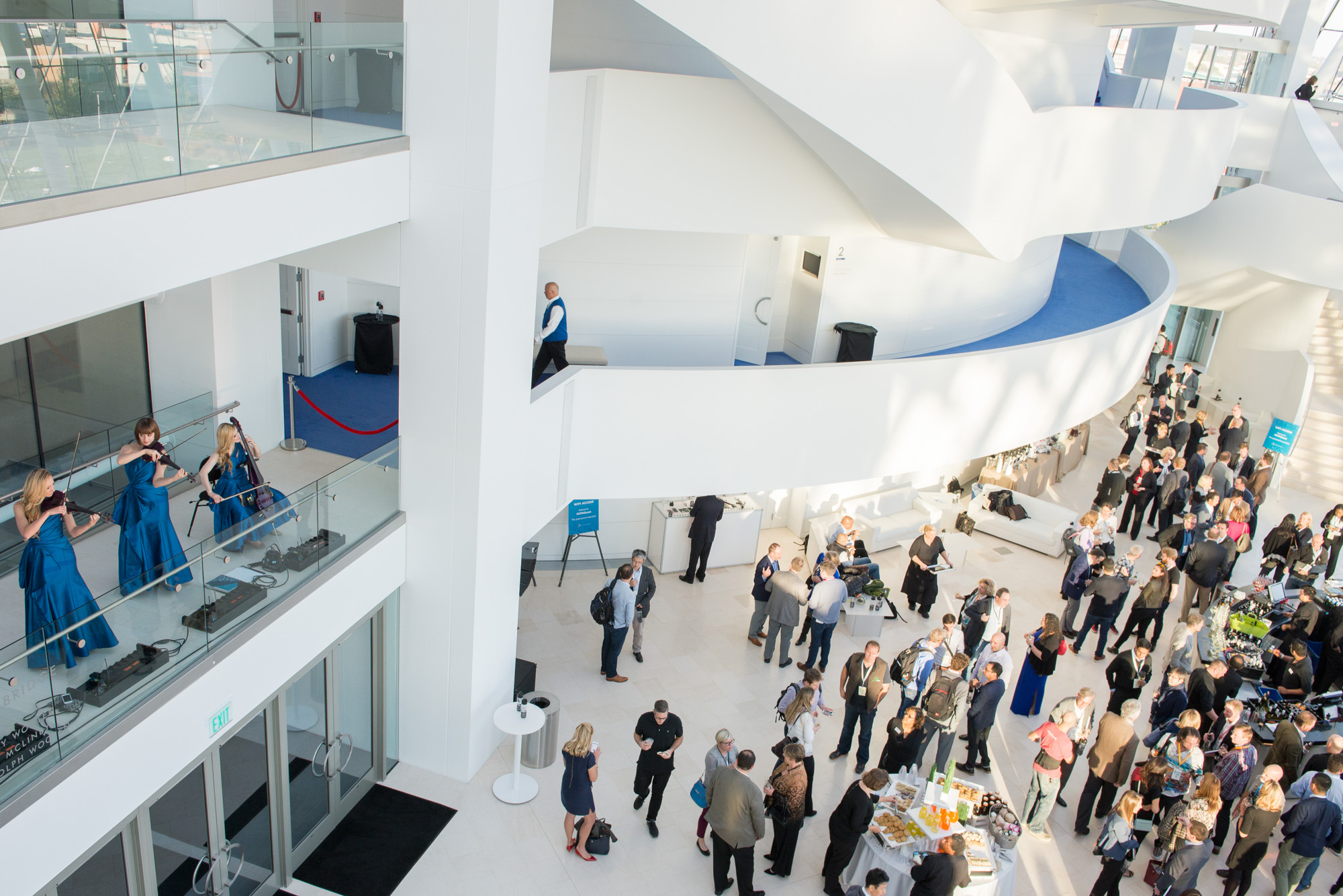 A group of people socializing in a spacious, modern indoor area with white architectural elements and a balcony where musicians from A Guide to the Kauffman Center are playing string instruments.