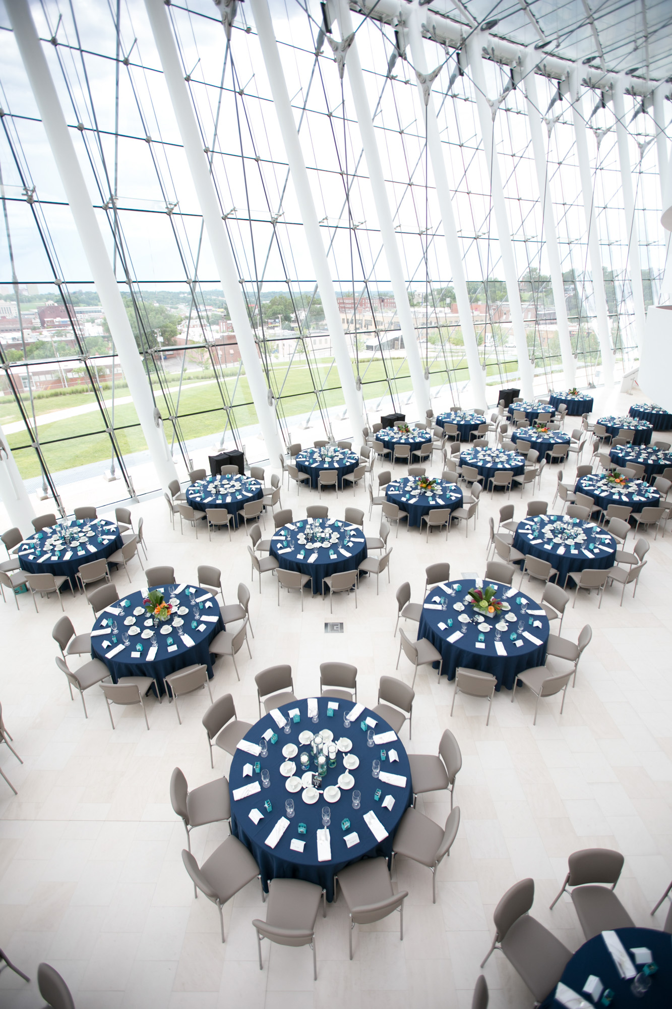 A spacious event hall with large windows showcases exquisite architecture, featuring round tables covered with navy blue tablecloths and beige chairs arranged around them. The setup is perfect for a formal gathering in such elegant spaces.