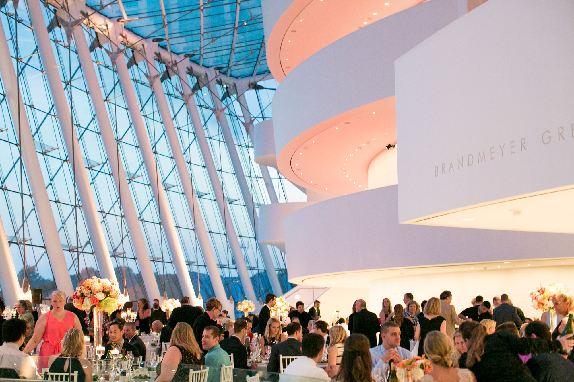 A crowd of people are seated at tables, attending an event in a modern, spacious venue with large curved structures and tall windows in the background, where several corporate sponsorships are prominently featured.