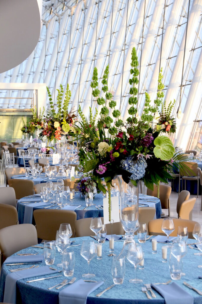 A decorated dining area with tables covered in blue tablecloths, set with glassware and floral centerpieces, is situated within a spacious, modern venue with large, slanted windows—perfect for those looking to book through our Special Event Rental Form.