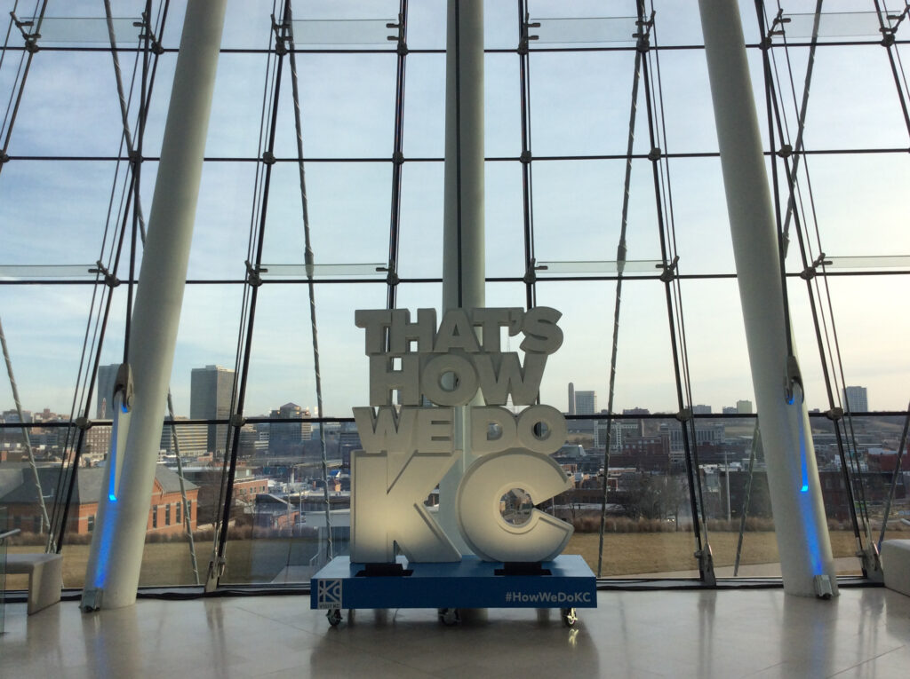 View from inside a modern building with large windows, showing a city skyline. A large sign reads "THAT'S HOW WE DO KC" with "#HowWeDoKC" on the base.