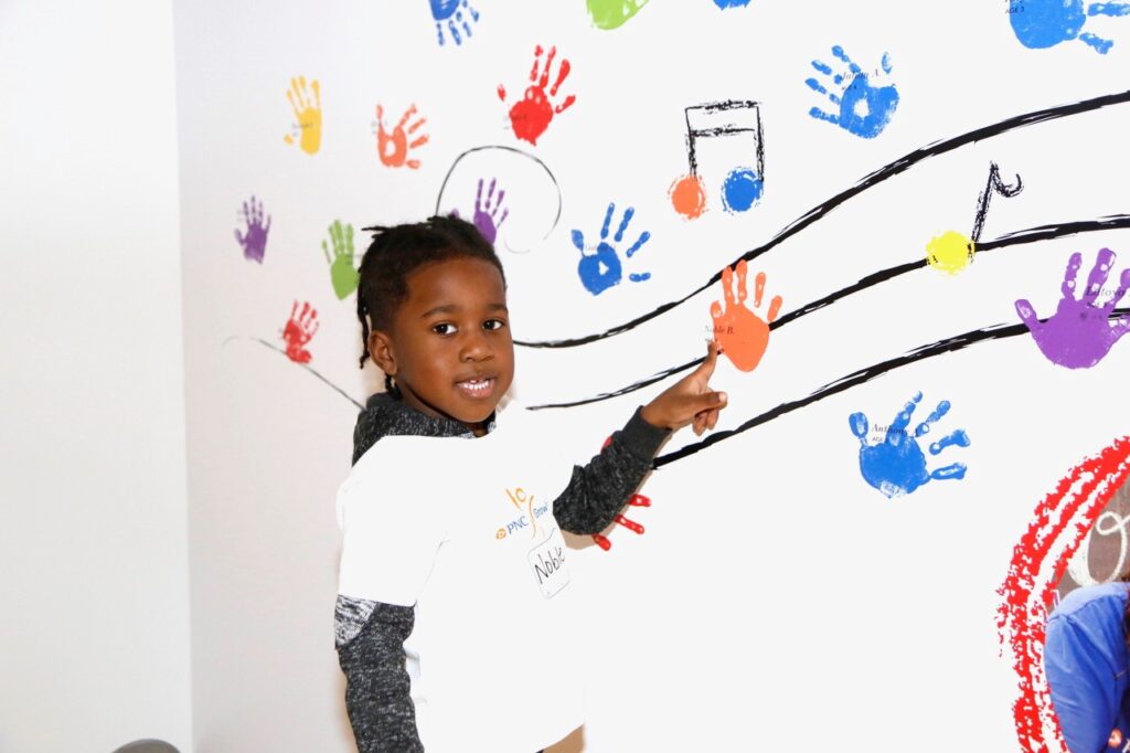 A child points to a colorful mural on a wall featuring musical notes and handprints in various colors, wearing a white shirt.