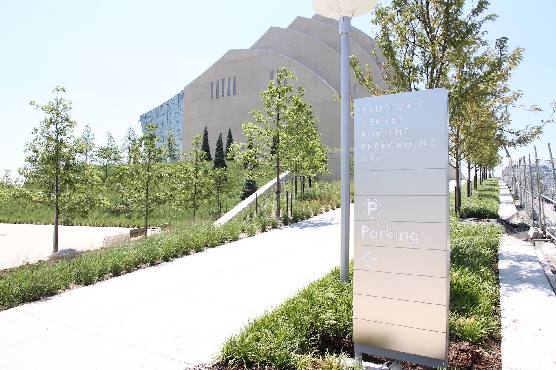 The image shows the Kauffman Center for the Performing Arts with a sign indicating directions & parking, surrounded by young trees and manicured grass.