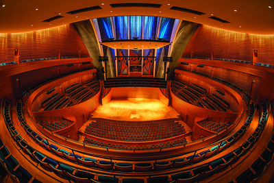 A wide-angle view of an empty, modern concert hall featuring circular seating arrangements, a wooden stage, and blue-lit ceiling panels.