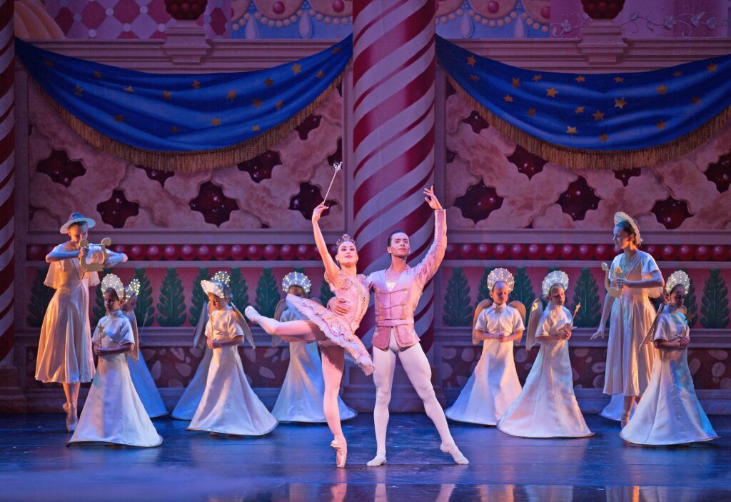 Ballet dancers perform on stage in ornate costumes, with a pink-decorated backdrop. A female dancer balances on one leg, while a male dancer stands beside her. This captivating scene feels like a highlight from the Performing Arts Sampler, with other dancers gracefully poised in the background.