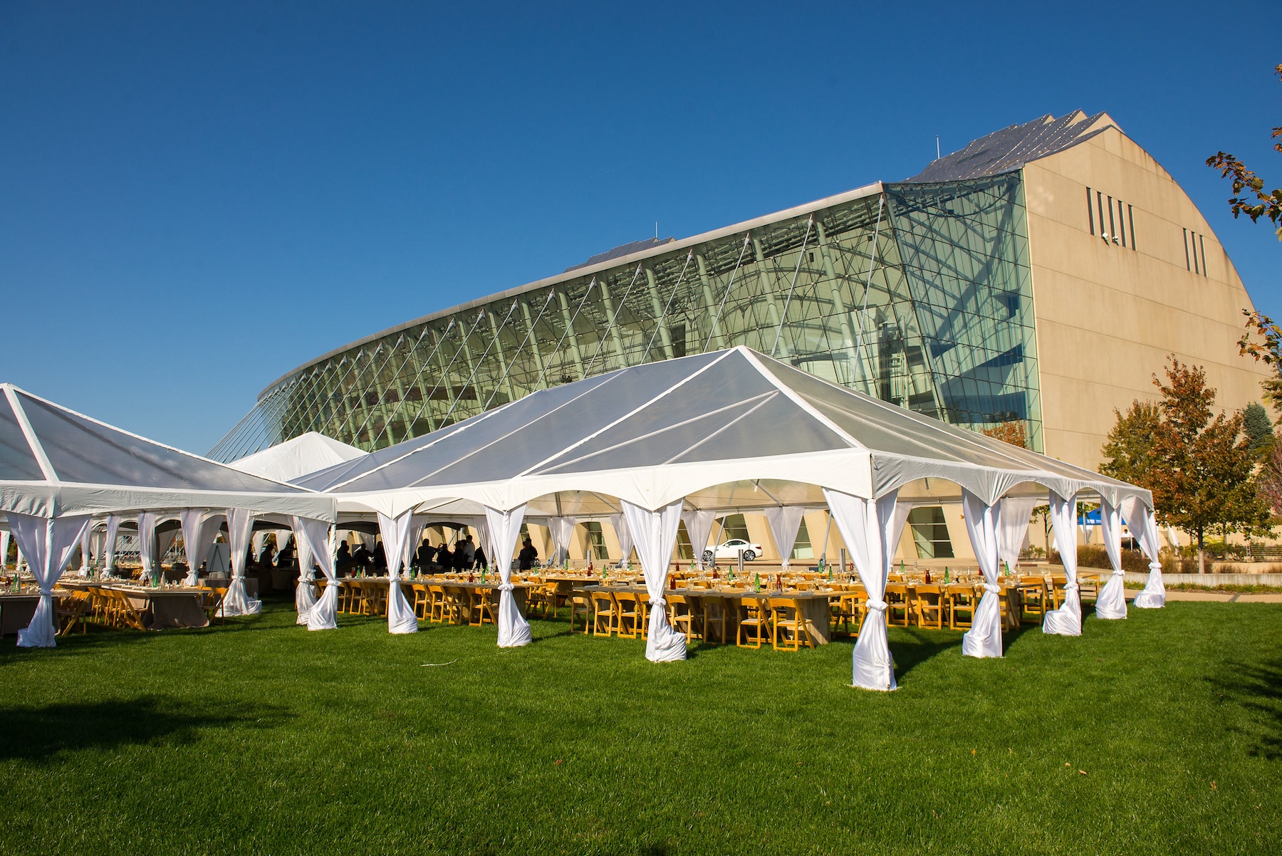 A large, white event tent set up on a green lawn in front of the modern glass and concrete Kauffman Center under a clear blue sky. Rent this perfect setting for your next outdoor occasion.