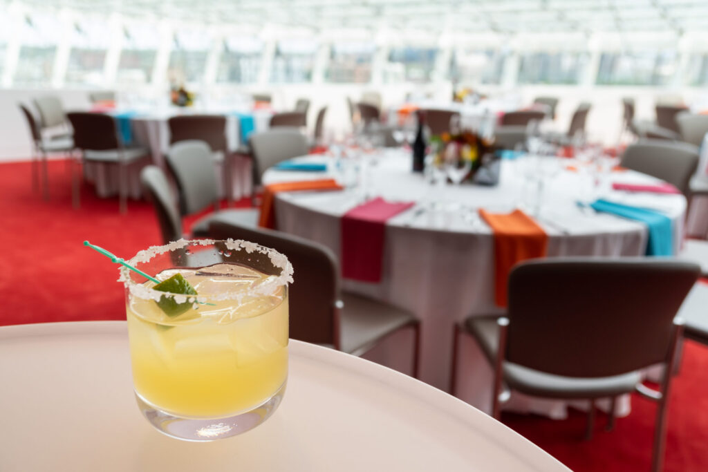 A cocktail garnished with a lime and salt rim sits on a table in focus, with blurred background of round tables set up for an event.