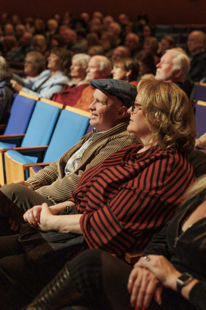 A man and a woman sit in a crowded auditorium, attentively watching a performance or presentation.