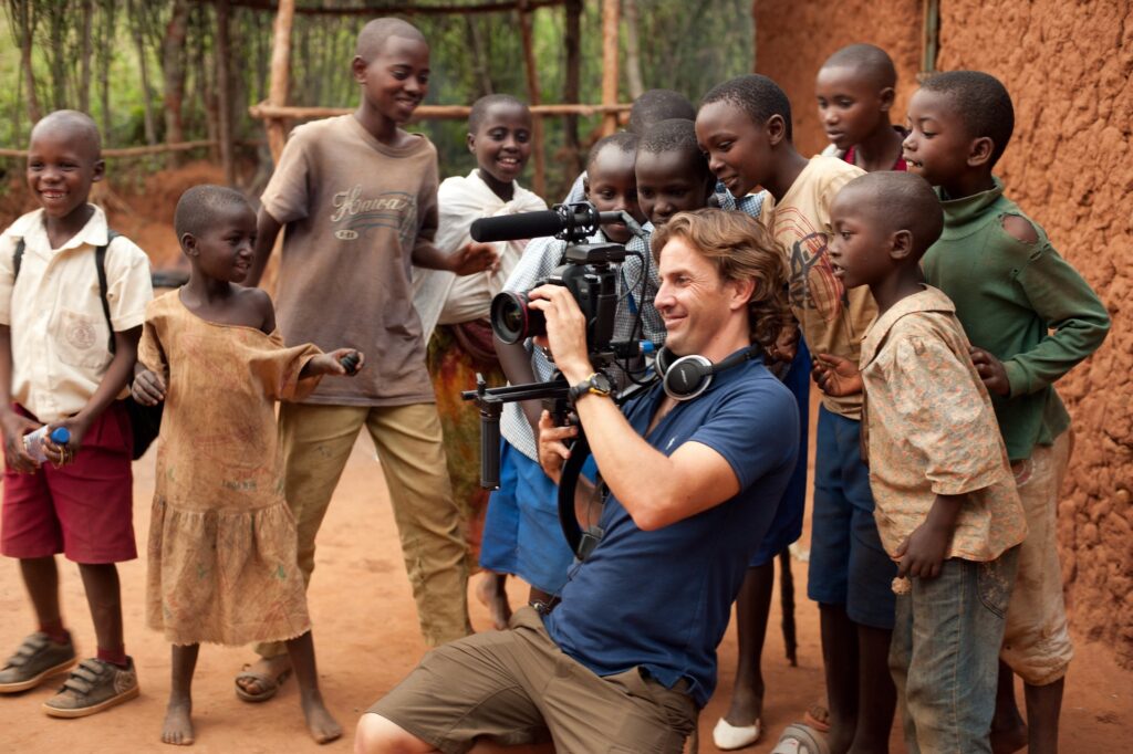 A man with a camera films while smiling children gather around him in an outdoor setting with dirt ground and mud-brick buildings, capturing the joy of nature.