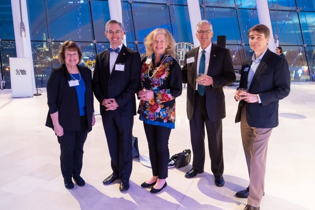 A group of five people in formal attire stand together, smiling, in a modern, glass-walled venue. They appear to be at a social or professional event celebrating business memberships.