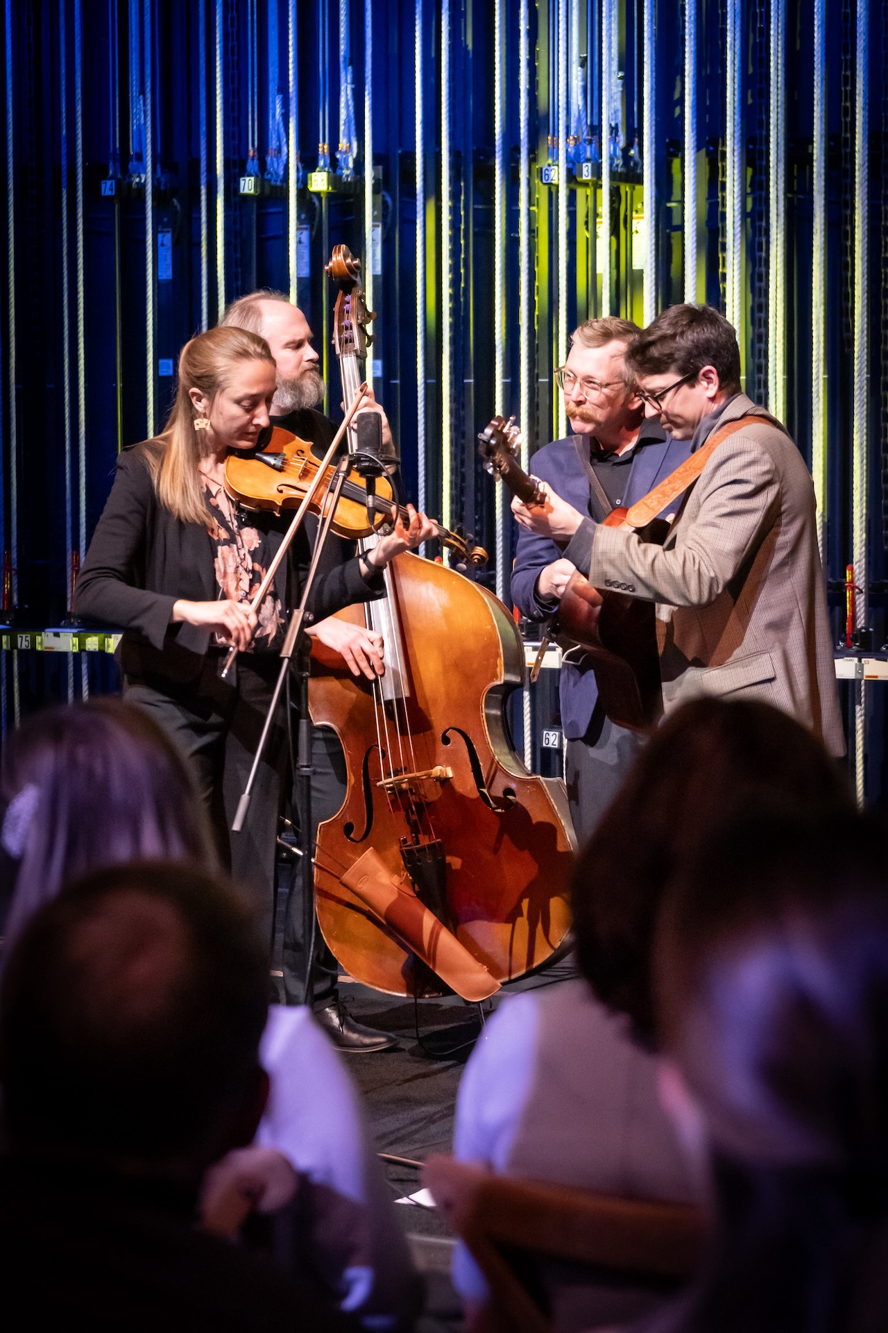 Four musicians play string instruments on stage, including a violin, double bass, and two guitars. The audience, many of whom hold business memberships, watches them perform.