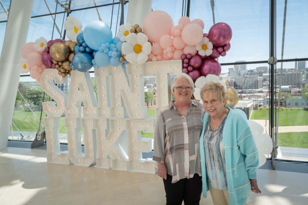 Two women stand smiling in front of a "SAINT LUKE" sign with balloons, inside a modern glass building with a city view in the background.