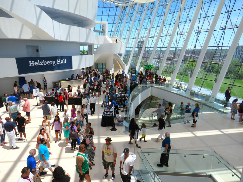 A large crowd gathered in a modern, sunlit atrium with glass walls and a "Helzberg Hall" sign, buzzing with excitement at Kauffman Center